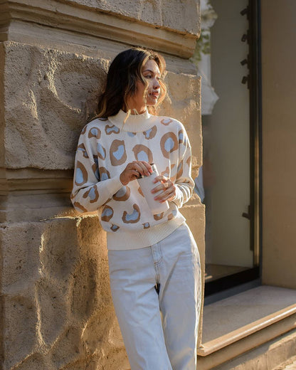 Woman wearing a patterned sweater and white pants standing against a stone wall.