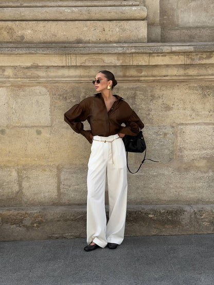 Woman in brown shirt and white pants standing against a stone wall.