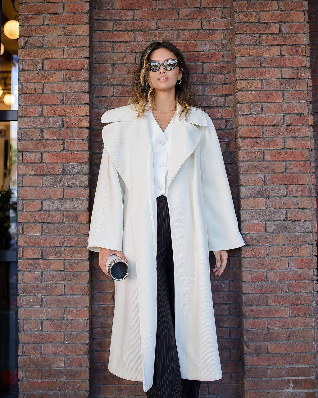 Woman wearing a long white coat against a brick wall.