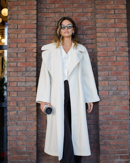 Woman wearing a long white coat against a brick wall.