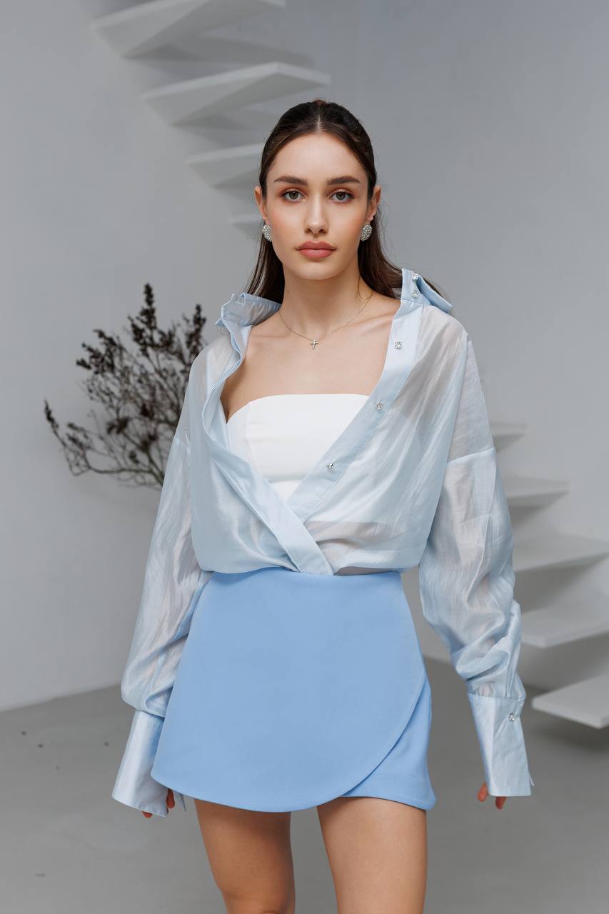 Woman wearing a light blue outfit with a white top and skirt, standing against a neutral background.