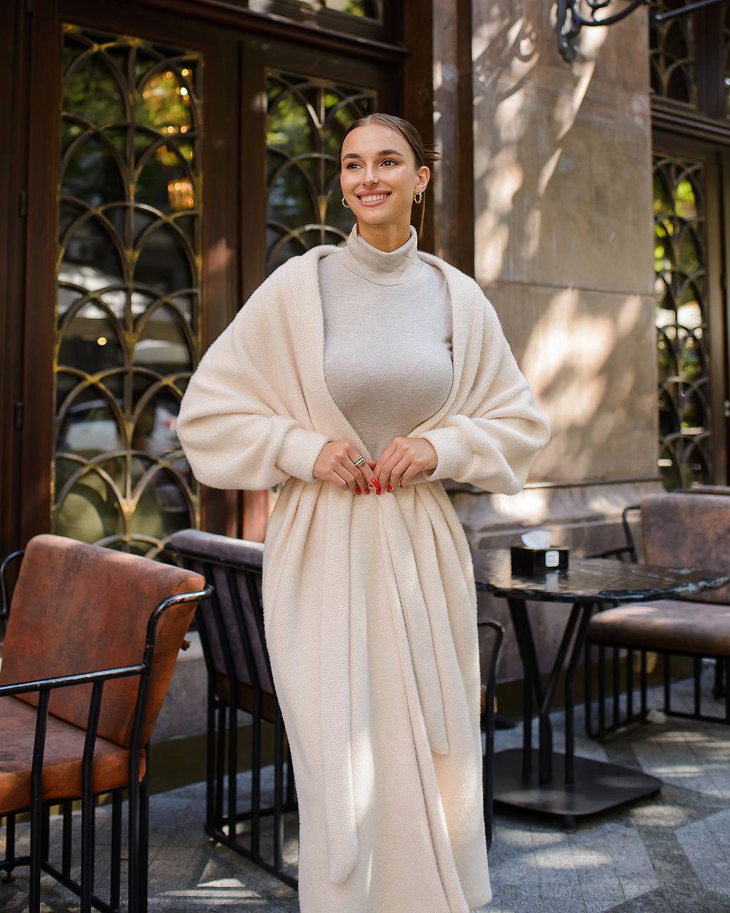 Woman in a long beige cardigan standing outdoors in a cafe setting
