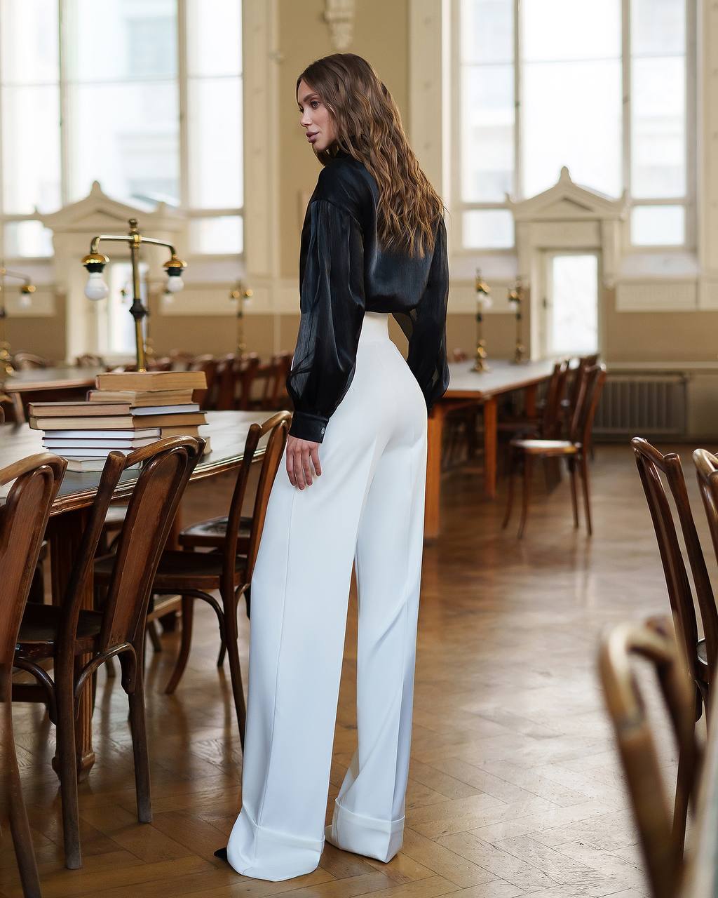 Woman in black top and white pants standing in a large room with wooden tables and chairs.