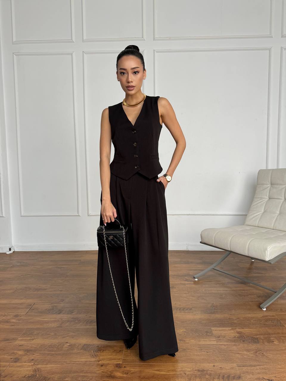 Woman in a black sleeveless top and wide-leg pants holding a black handbag against a white paneled wall.