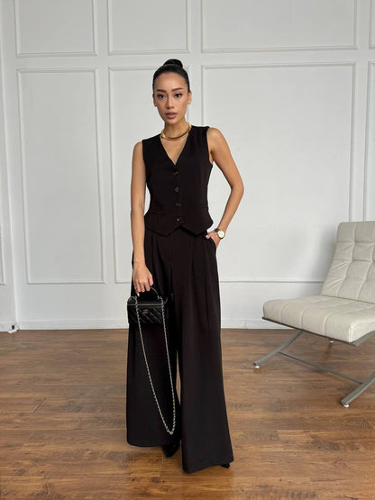 Woman in a black sleeveless top and wide-leg pants holding a black handbag against a white paneled wall.