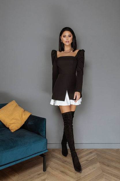 Woman in a black dress with white trim and knee-high boots standing in a room with a gray wall and blue couch.