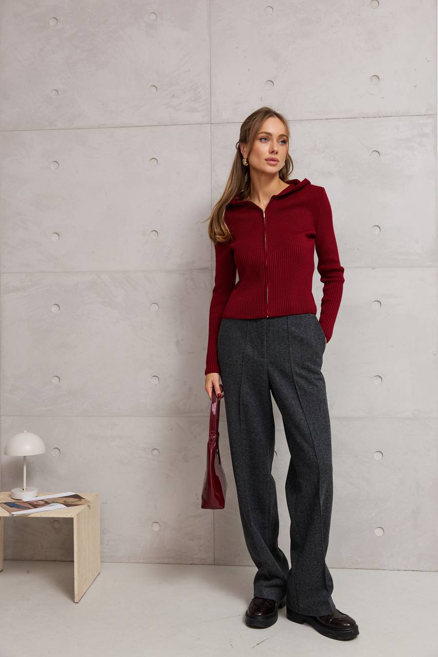 Woman in a red sweater and gray pants standing against a light gray wall.