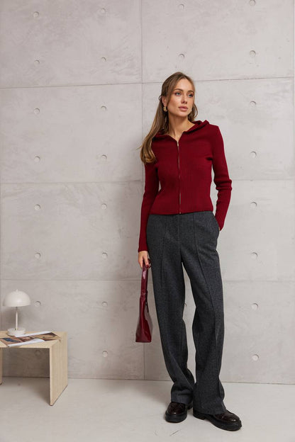 Woman in a red sweater and gray pants standing against a light gray wall.