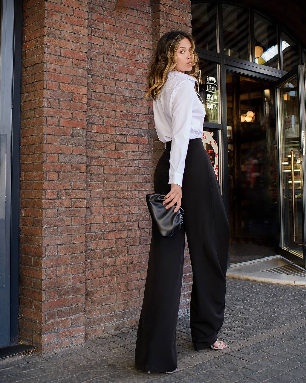 Woman in a white blouse and black pants standing against a brick wall.