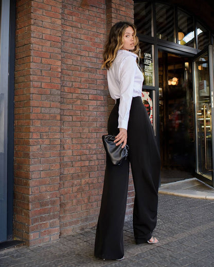 Woman in a white blouse and black pants standing against a brick wall.