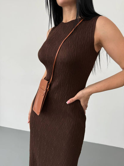 Woman wearing a brown sleeveless dress with a brown leather bag over her shoulder against a white background.