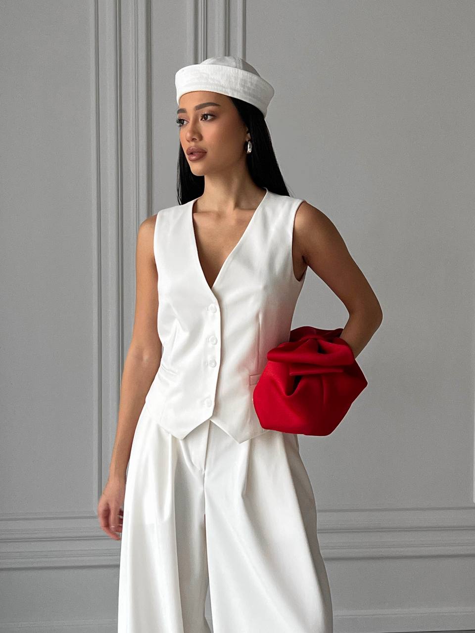 Woman in a white outfit holding a red clutch against a gray wall