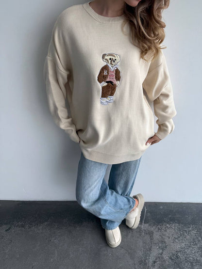 Person wearing a beige sweater with a graphic design of a bear and text, standing against a plain background.