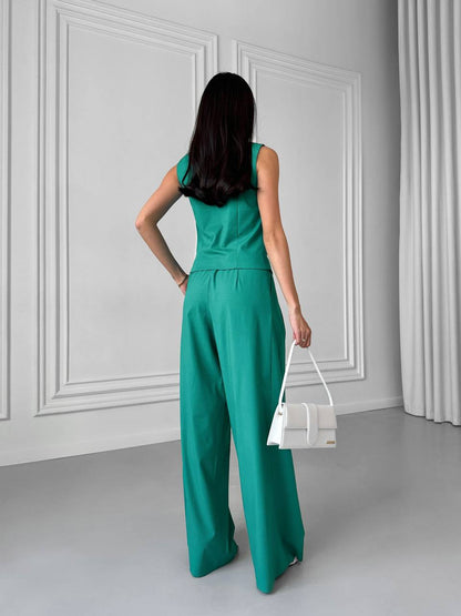 Woman wearing a teal jumpsuit holding a white handbag in a minimalistic room.