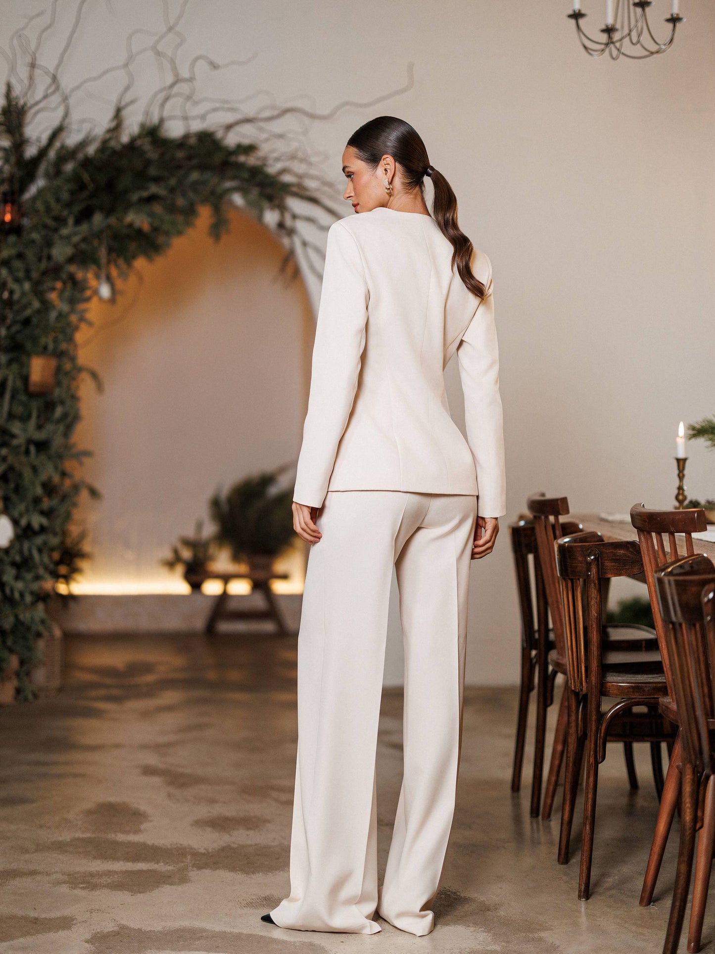 Woman in a white suit standing in a decorated room with plants and wooden furniture.