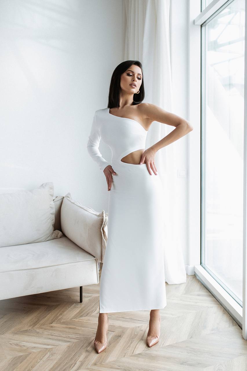 Woman in a white one-shoulder dress standing in a bright room with large windows.