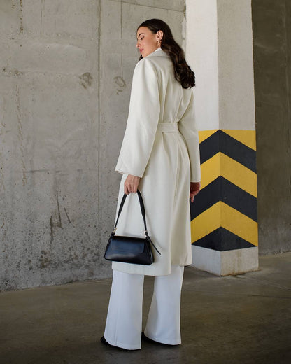 Woman in a long beige coat holding a black handbag against a concrete wall with a yellow and black zigzag pattern.