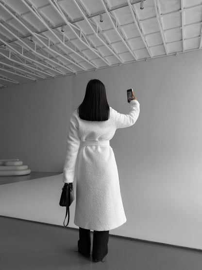 Person in a white coat taking a selfie in a minimalistic room.