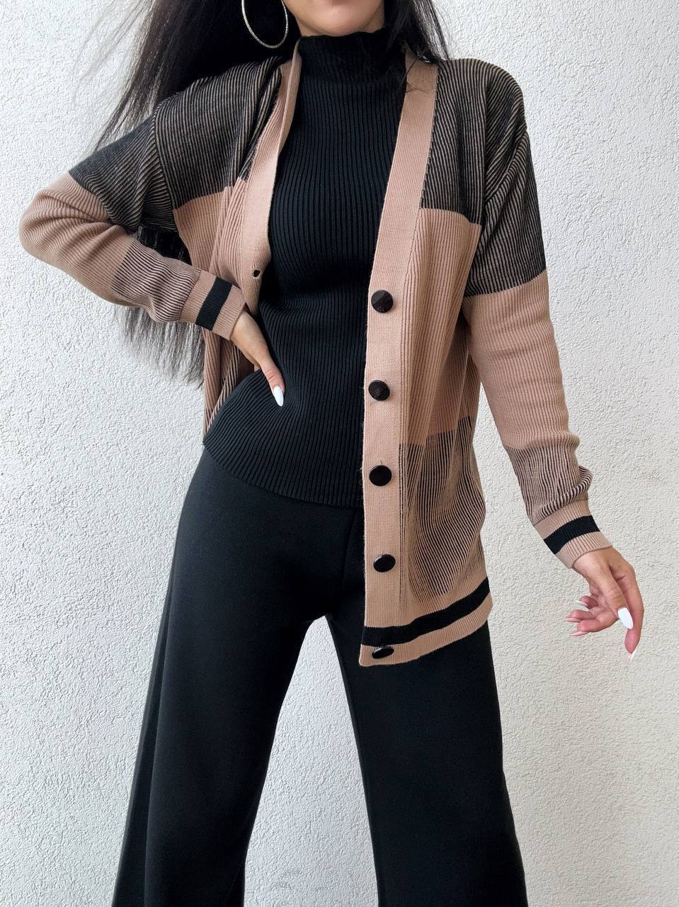 Person wearing a long beige cardigan with black buttons over a black outfit against a light background