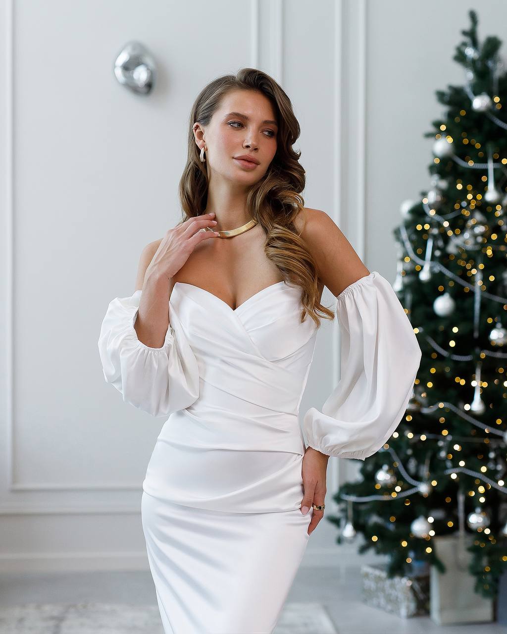 Woman in a white off-shoulder dress standing in front of a decorated Christmas tree.