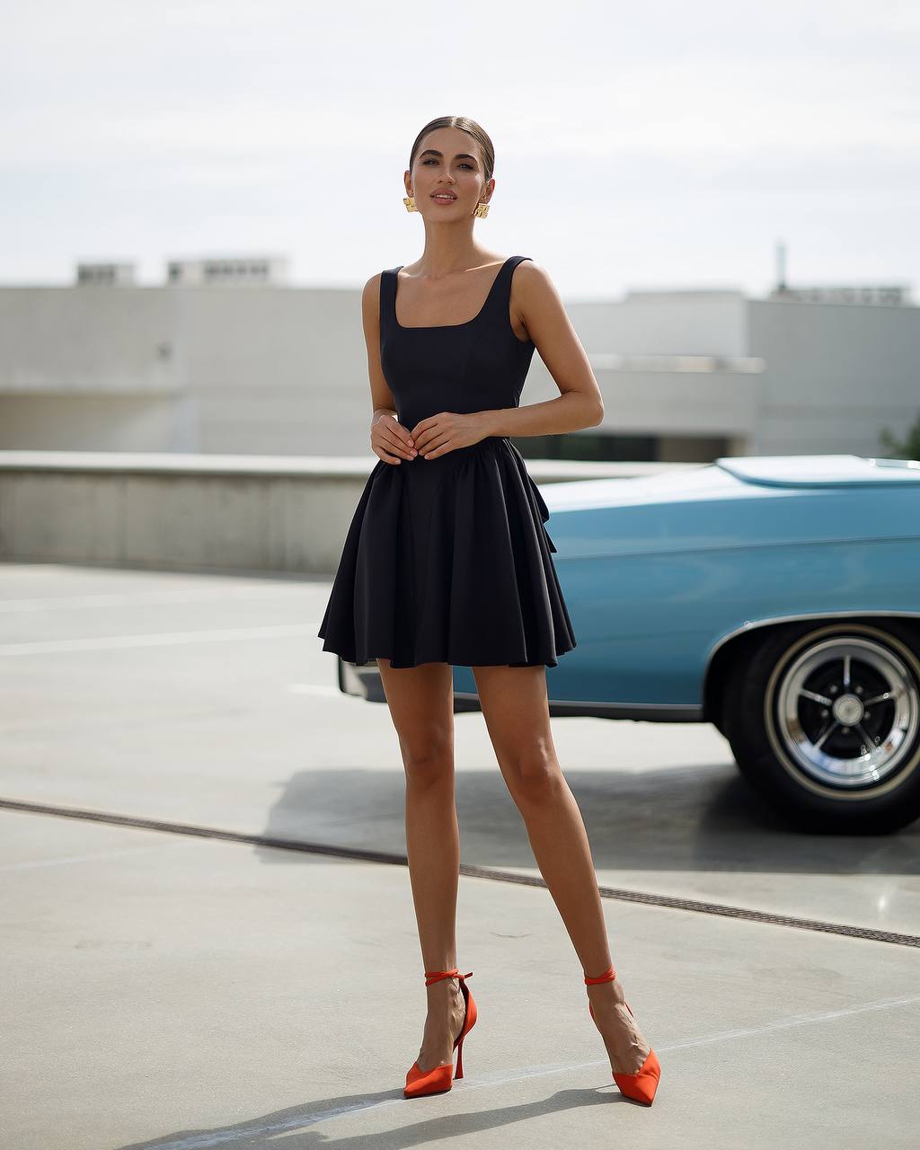 Woman in a black dress with orange shoes standing in front of a blue car.