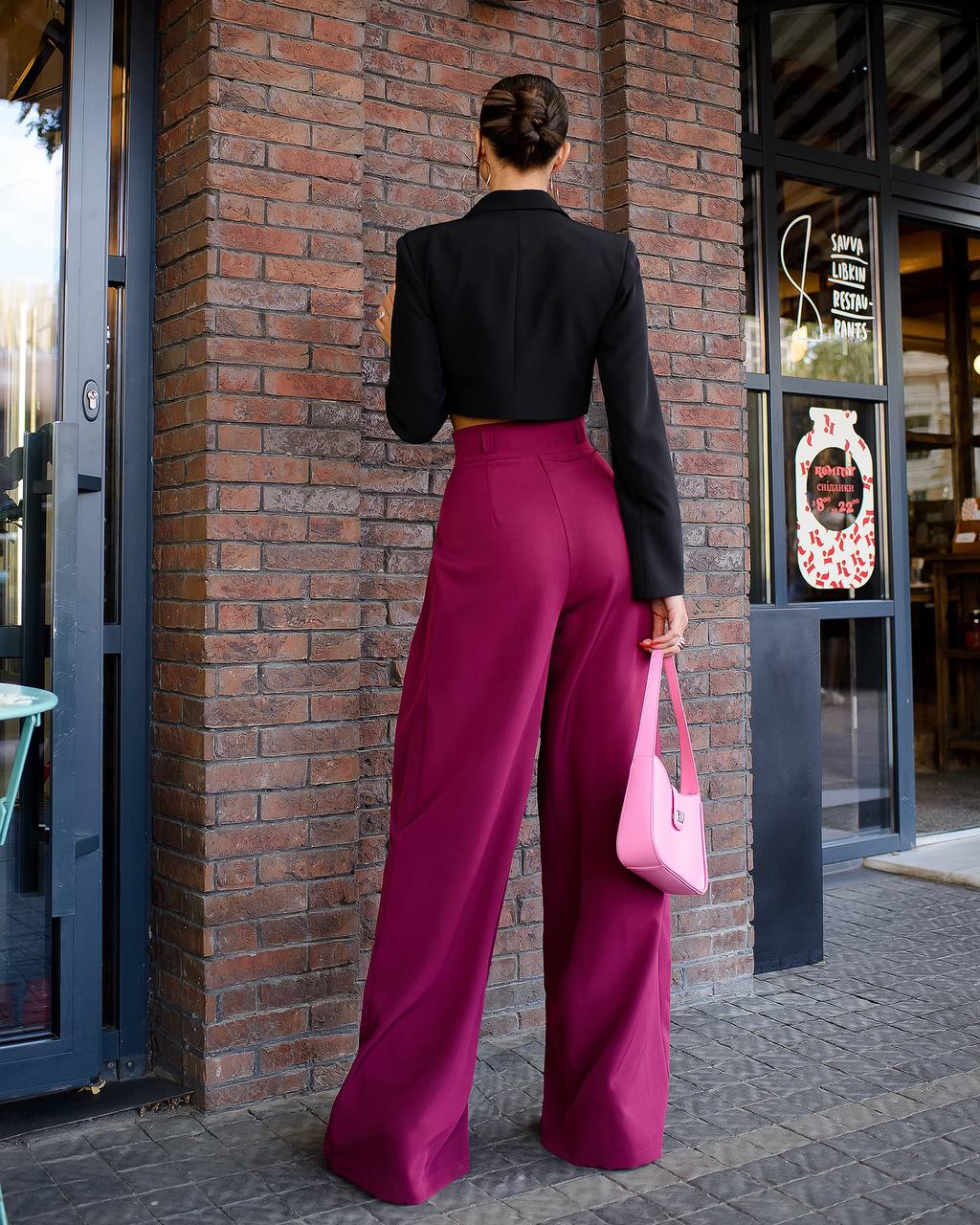 Person wearing a black top and magenta pants holding a pink bag against a brick wall.