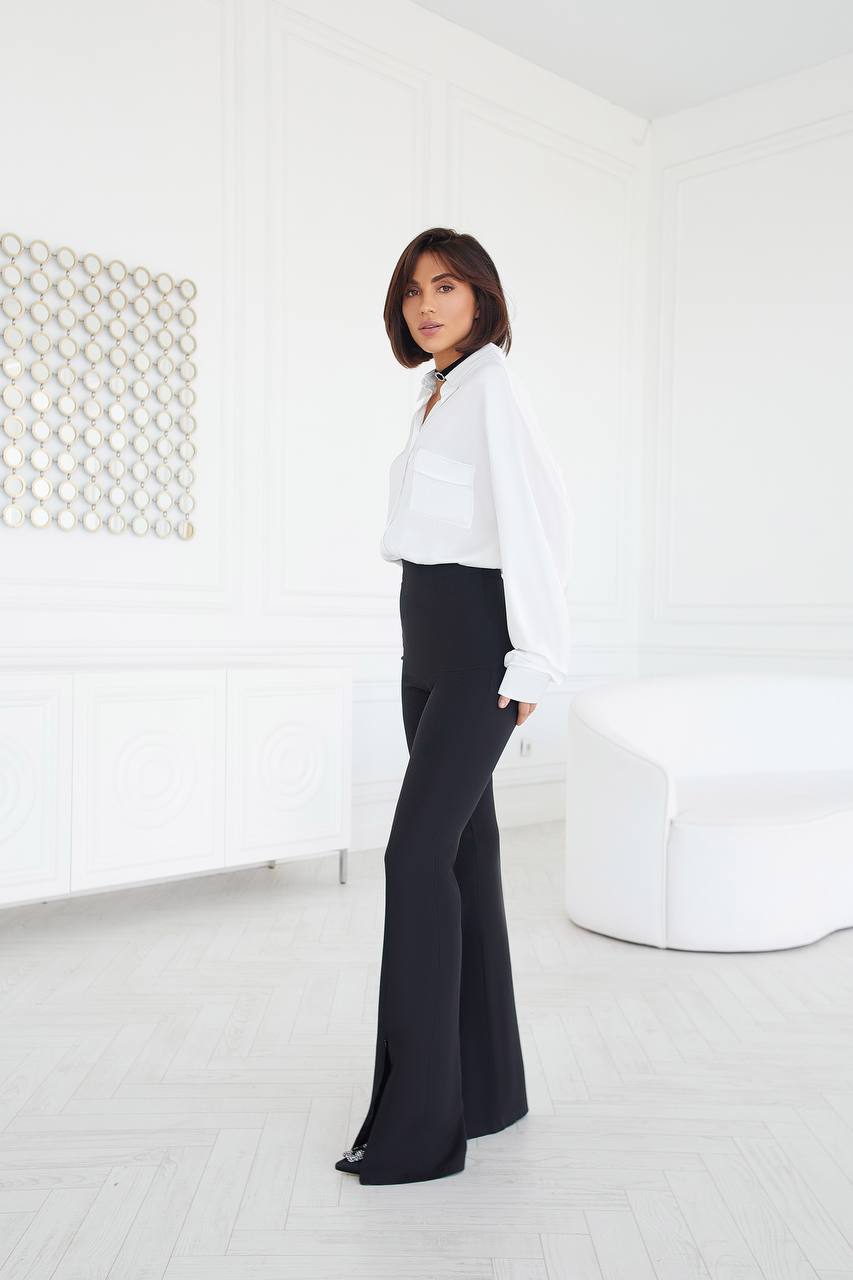 Person wearing a white blouse and black pants in a minimalistic room.