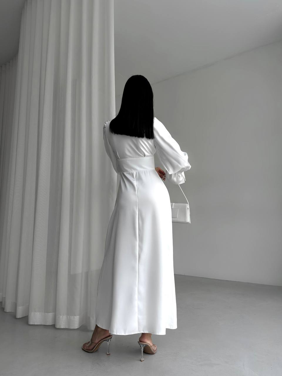 Woman in a long white dress standing in a minimalistic room with a curtain.