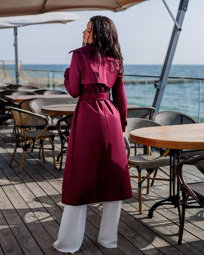 Person wearing a long burgundy coat standing on a wooden deck with tables and chairs, looking out towards the ocean.