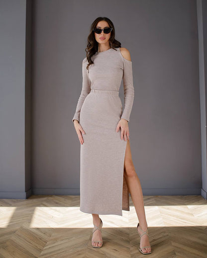 Woman wearing a long, light-colored dress with a slit in a room with gray walls and wooden flooring.