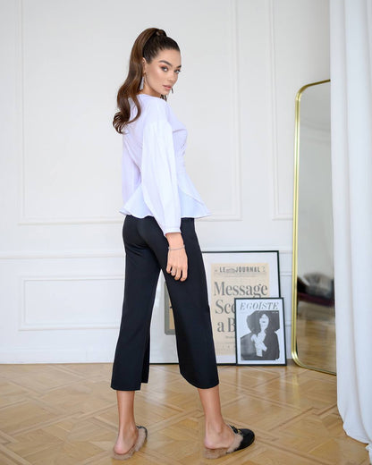 Woman wearing a white blouse and black pants in a room with a mirror and framed picture on the wall.