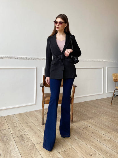 Woman in a black blazer and blue flared pants standing in a room with wooden flooring and white walls.