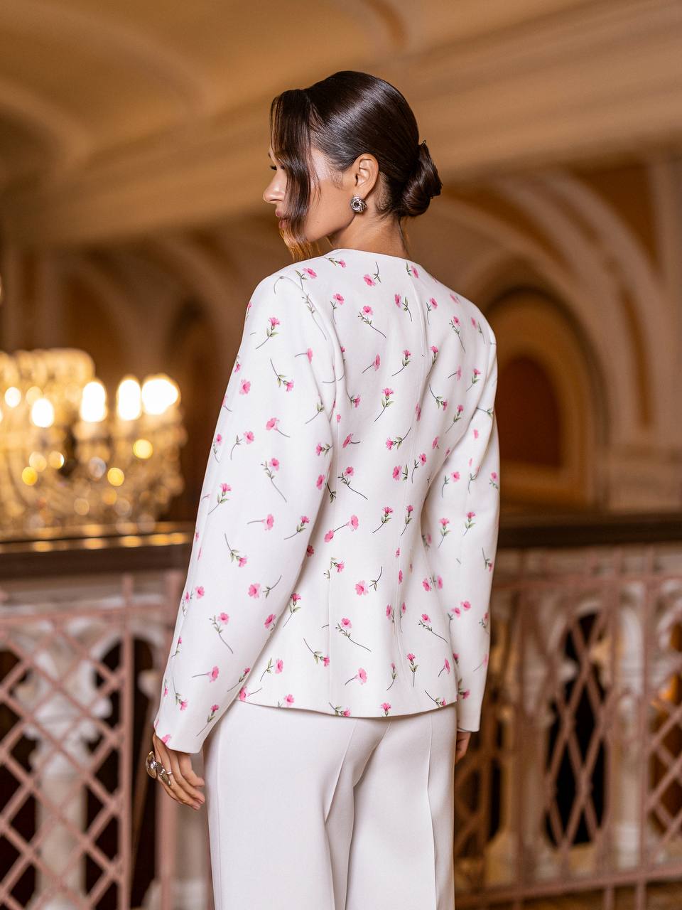 Woman in a white floral blouse and pants standing in an elegant indoor setting.