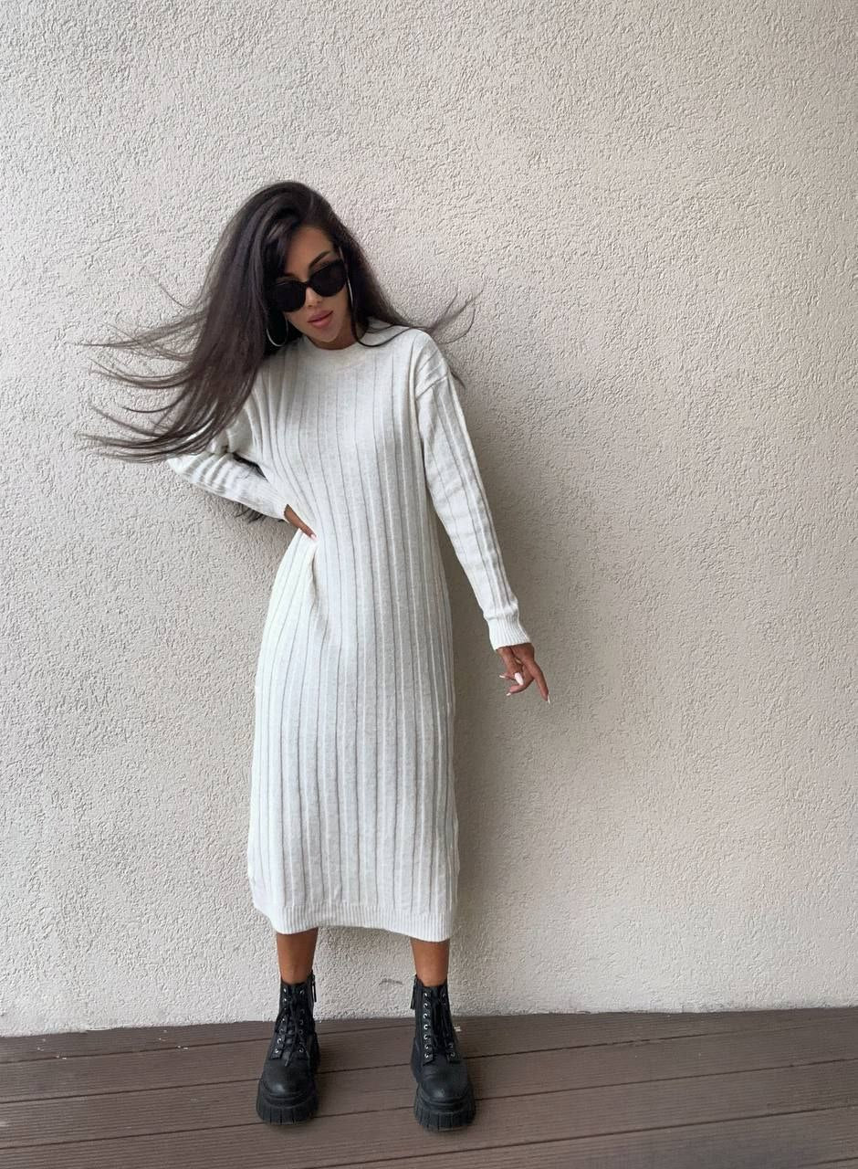 Woman wearing a white ribbed dress with black boots against a plain wall.