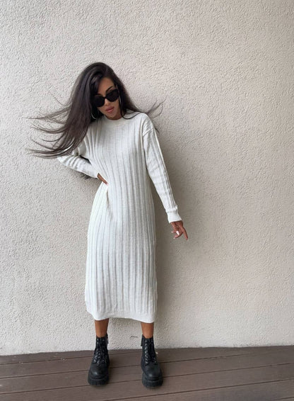 Woman wearing a white ribbed dress with black boots against a plain wall.