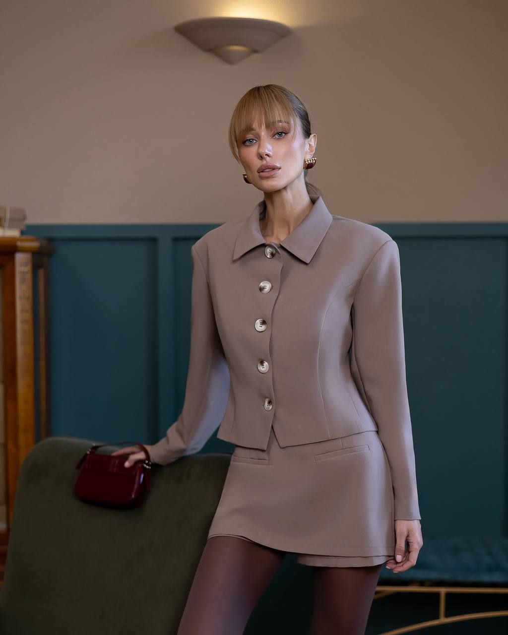 Woman in a beige suit holding a red handbag in an indoor setting