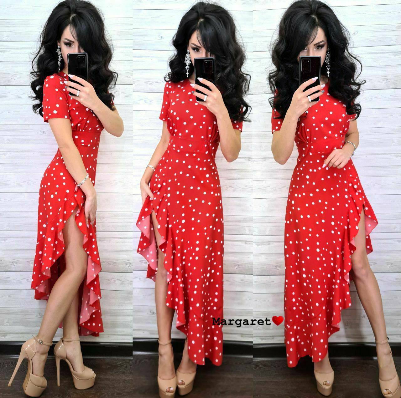 Woman wearing a red polka dot dress with a slit, posing in front of a white brick wall.
