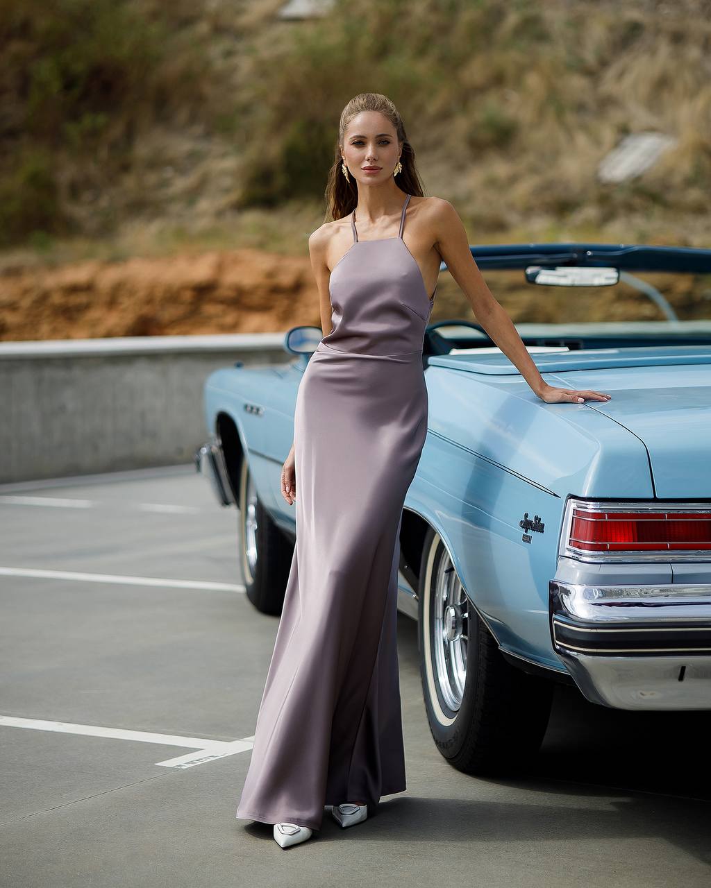 Woman in a long purple dress standing next to a classic blue car on a road.