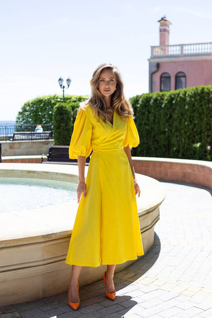 Woman in a yellow dress standing outdoors near a fountain with greenery and a building in the background.