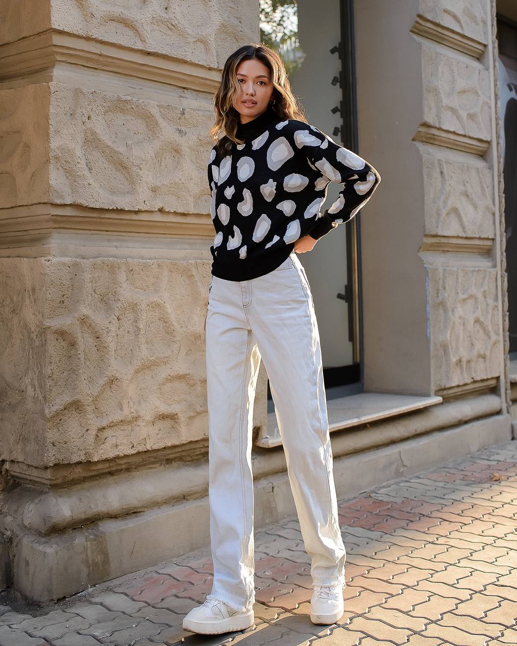 Woman wearing a black sweater with white heart patterns and white pants standing in front of a stone building.