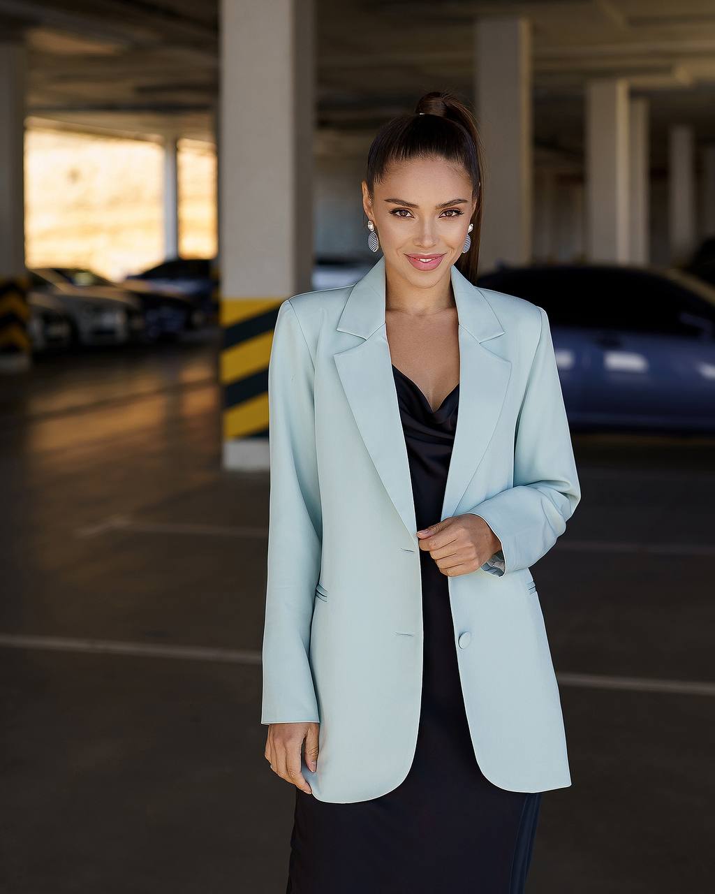 A woman stands in a parking garage wearing a light blue tailored blazer over a black dress or top. The background shows concrete pillars, parked cars, and soft natural light coming from the left.
