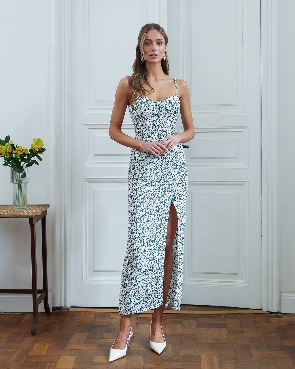 Woman in a floral dress standing in a room with wooden flooring and white walls.