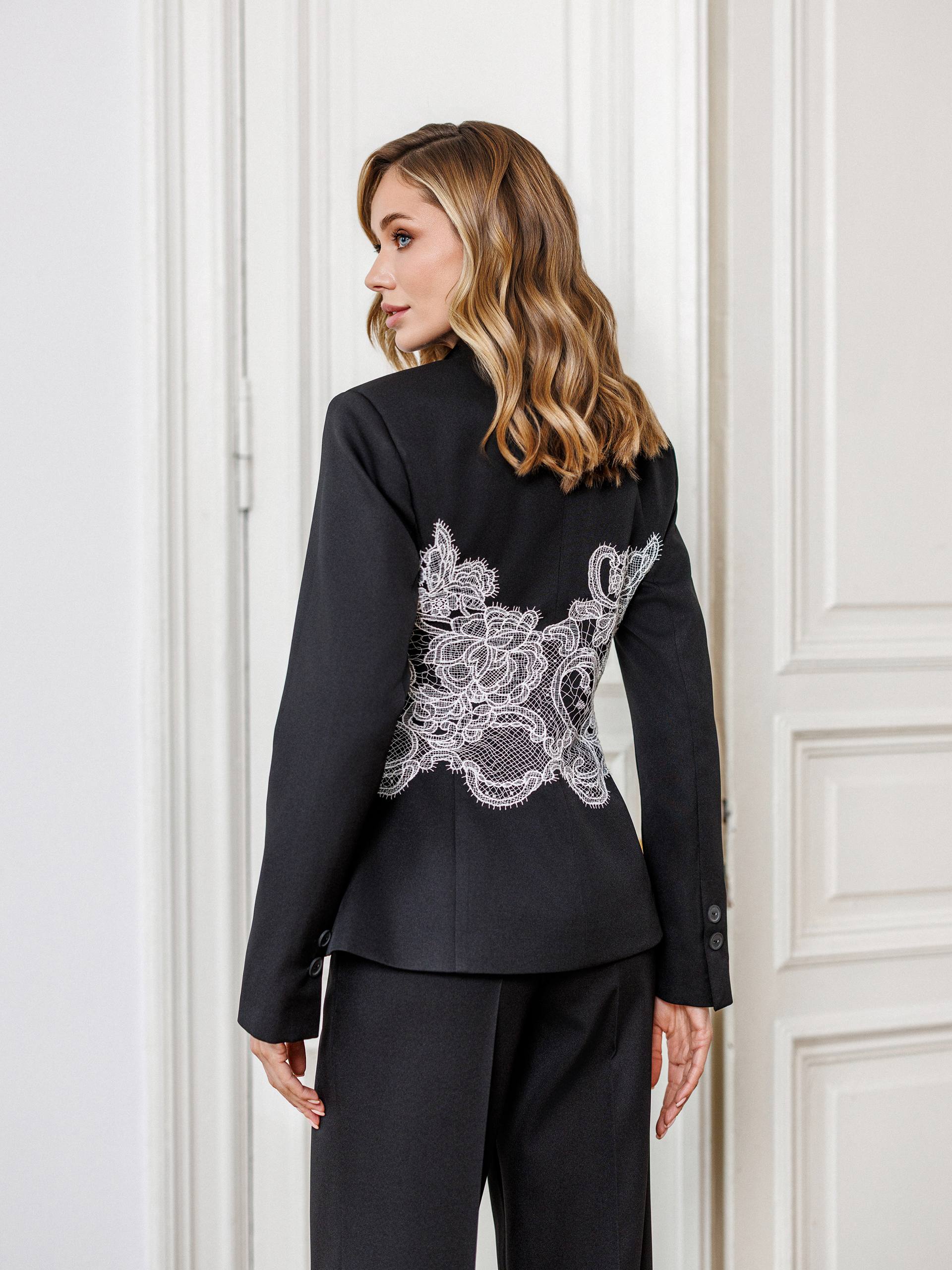Woman wearing a black suit with lace detailing on the back against a white wall.