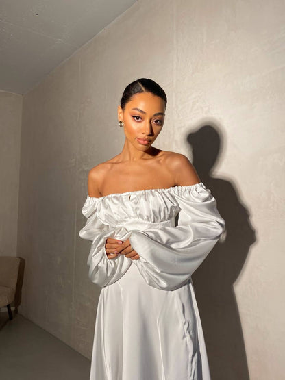 Woman wearing a white off-shoulder dress against a plain wall.
