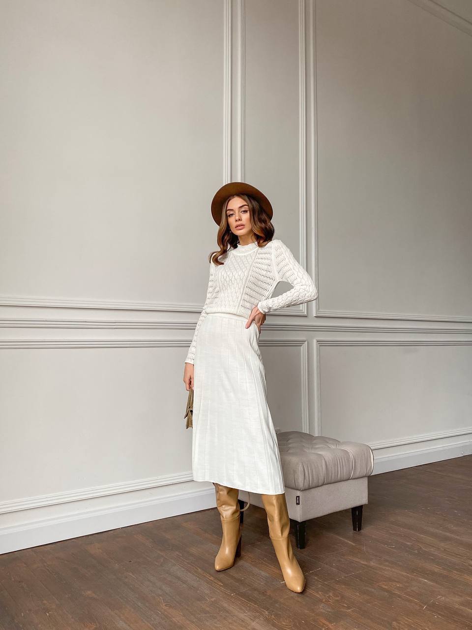 Woman in a white outfit with beige boots standing against a gray paneled wall.