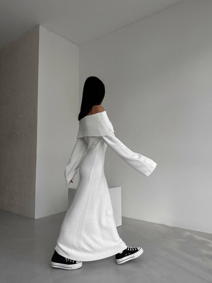 Person wearing a white off-shoulder sweater and black shoes in a minimalistic room.