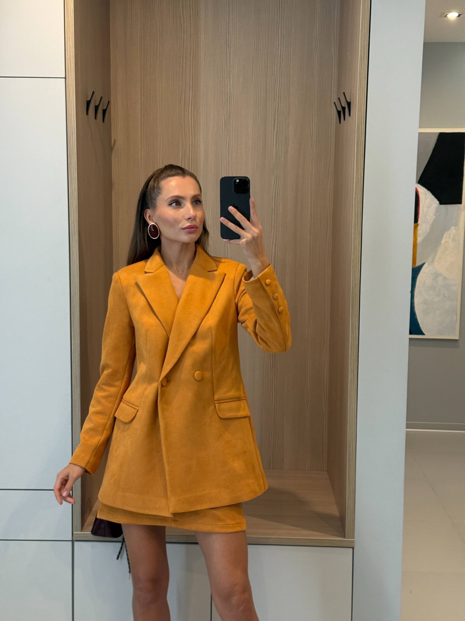 A woman takes a mirror selfie wearing a mustard yellow tailored blazer and matching mini skirt set in a modern indoor setting with light wood and white paneling.