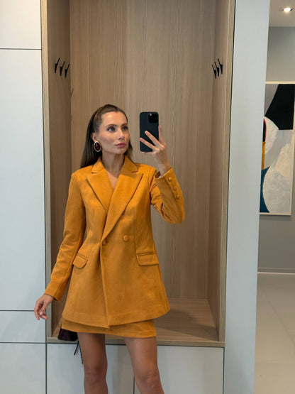A woman takes a mirror selfie wearing a mustard yellow tailored blazer and matching mini skirt set in a modern indoor setting with light wood and white paneling.