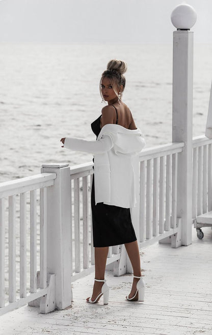 Woman in black dress and white jacket standing on a white wooden deck with a white wall background
