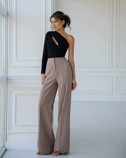 Woman wearing a black top and beige pants against a white paneled wall.